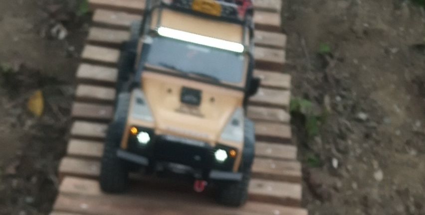Eine Holzrampe führt von einem kleinen Hügel hinab. Ein Spielzeug-Jeep mit Frachtablage fährt darauf herunter.