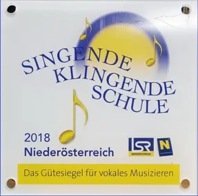 Ein weißes Schild mit blauem und gelbem Text zeigt 'Singende Klingende Schule 2018 Niederosterreich'. Es enthält einen gelben Musiknote. Das gelbe Banner liest 'Das Gütesiegel für vokales Musizieren'. Logos für 'ISR' und 'N' sind ebenfalls vorhanden.