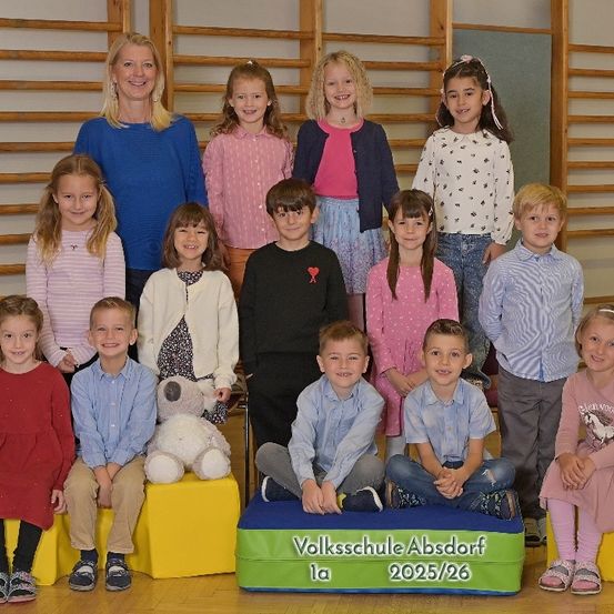 Gruppenfoto von Grundschulkindern und Lehrern, stehend und sitzend auf gelben Blöcken, lächelnd für die Kamera, mit einem Banner, das 'Volksschule Absdoorf 1a 2025/26' lautet.