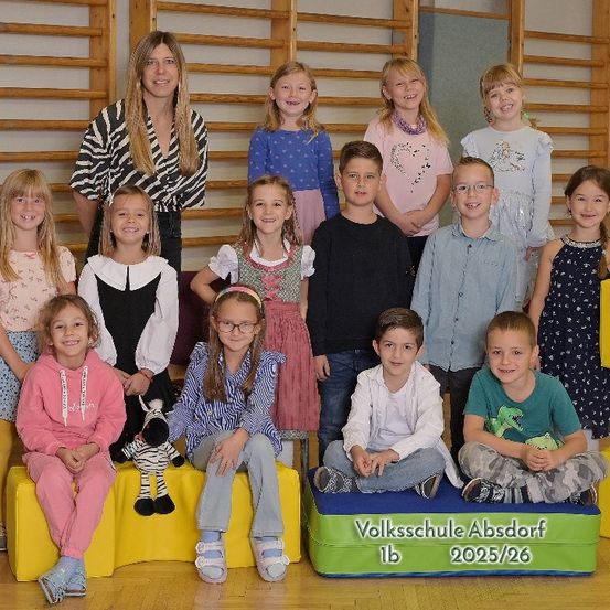 Gruppenfoto der 1b-Klasse der Volksschule Absdorf, aufgenommen im Jahr 2025/26. Die Schüler sitzen auf bunten Kissen, eine Frau steht hinter ihnen.