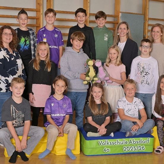 Eine Gruppe junger Schüler und Lehrer posiert für ein Foto, in Freizeitkleidung, einige halten ein Kuscheltier. Sie sitzen auf gelben Kissen, eines ist mit 'Volksschule Absdort 4a 2025/26' beschriftet. Im Hintergrund befinden sich Holzregale.