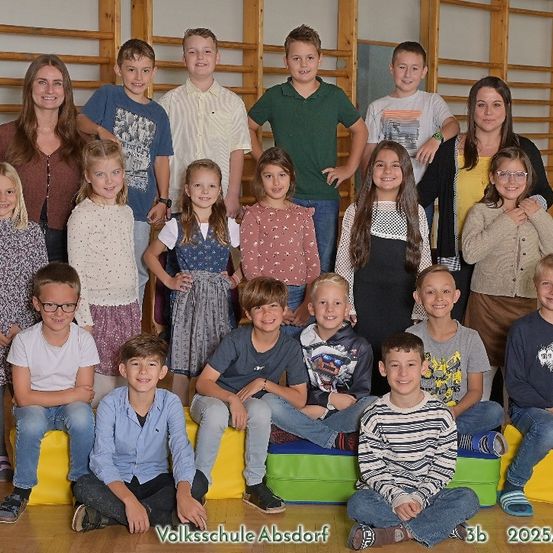 Gruppenfoto von Schülern und Lehrern der Volksschule Absdorf, aufgenommen in 2025/26. Die Kinder sitzen und stehen auf farbenfrohen Kissen, einige tragen Brillen und Turnschuhe.