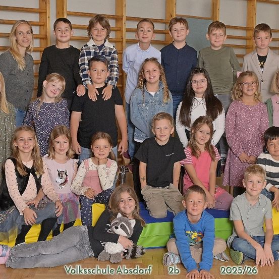 Eine Gruppe von Kindern und zwei Lehrern posiert für ein Foto in einer Turnhalle. Sie sitzen und stehen auf Kissen, mit einer Holzwand im Hintergrund.