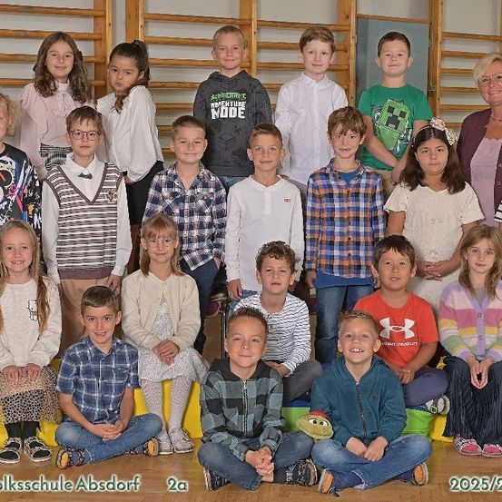 Eine Gruppenaufnahme von Schülern und einer Lehrerin an der Grundschule Absdorf, aufgenommen in 2025/26. Die Schüler sitzen und stehen, einige tragen Brillen und Turnschuhe, während die Lehrerin im Hintergrund steht.