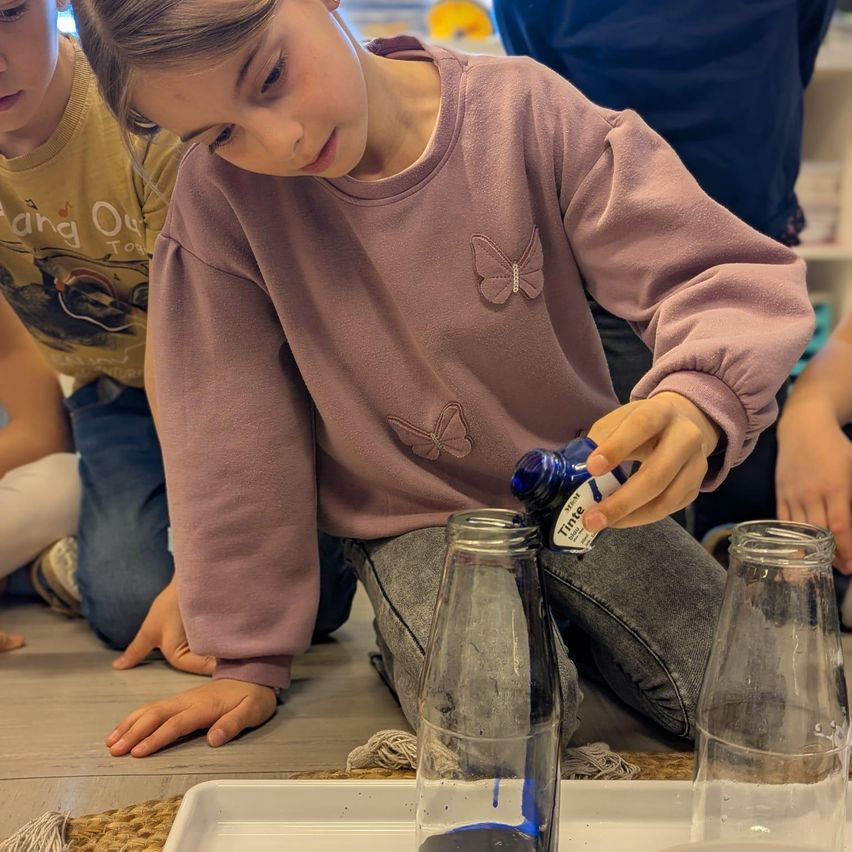 Ein junges Mädchen in einem rosa Sweatshirt gießt blaue Flüssigkeit in eine klare Glasflasche auf einem Tablett. Sie ist von anderen Kindern umgeben.