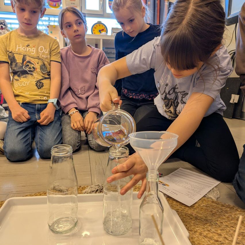 Eine Gruppe junger Kinder ist drinnen und beschäftigt sich mit einem wissenschaftlichen Experiment. Ein Kind gießt Wasser in einen Trichter. Es gibt leere Glasflaschen und ein weißes Tablett in der Nähe.