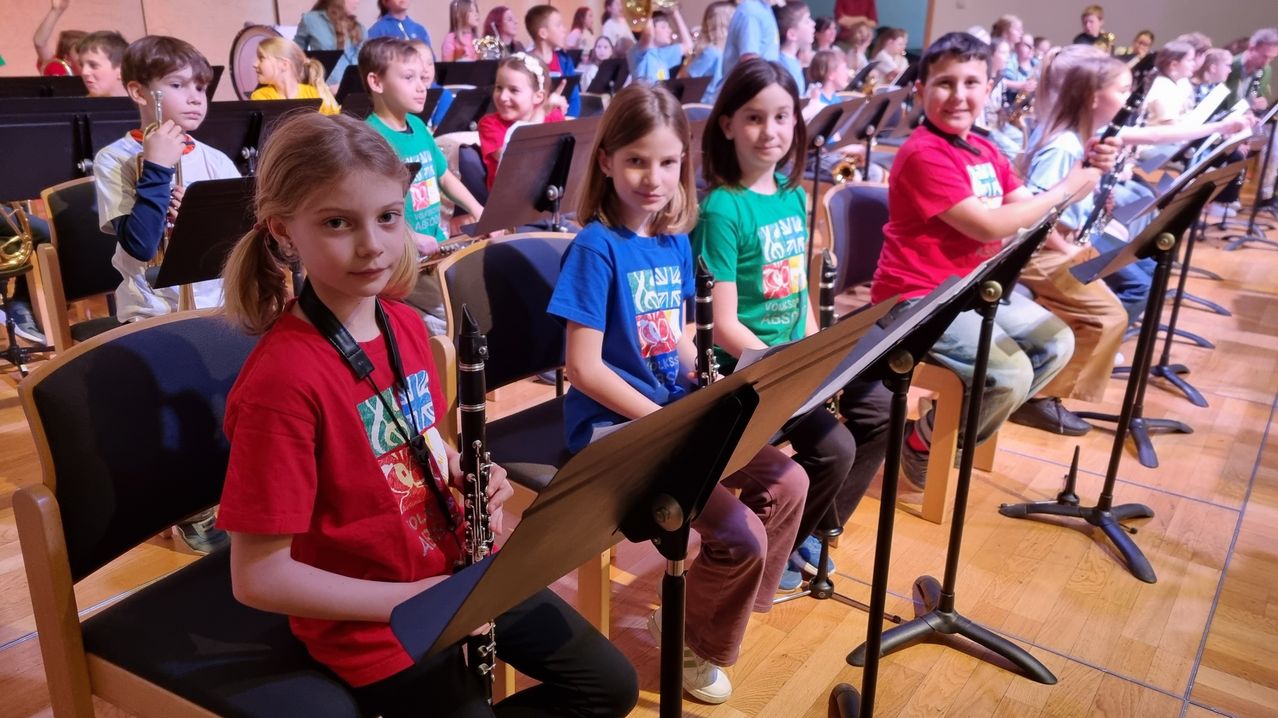 Mehrere Kinder sitzen während einer Bandprobe auf Stühlen. Sie haben Notenständer und Instrumente wie Klarinetten und Trompeten.