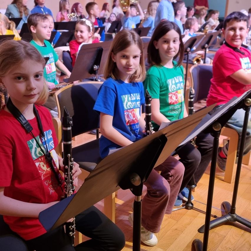 Mehrere Kinder sitzen während einer Bandprobe auf Stühlen. Sie haben Notenständer und Instrumente wie Klarinetten und Trompeten.