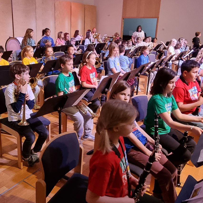 Ein Klassenzimmer voller Kinder, einige sitzend und andere stehend, halten Musikinstrumente in den Händen. Verschiedene Instrumente sind sichtbar, darunter Trompeten, Posaunen und Flöten. Die Kinder bereiten sich auf eine Musikstunde vor.