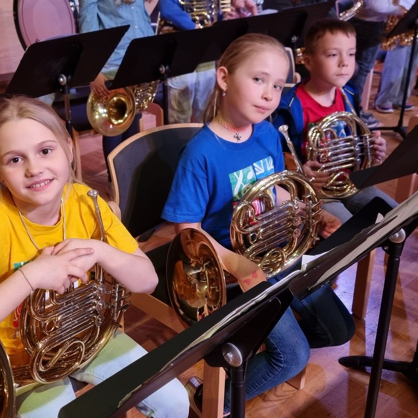 Drei Kinder, darunter ein Mädchen in Gelb und zwei Jungen in Blau, sitzen in Stühlen und halten französische Hörner, mit Notenblättern und anderen Instrumenten im Hintergrund.