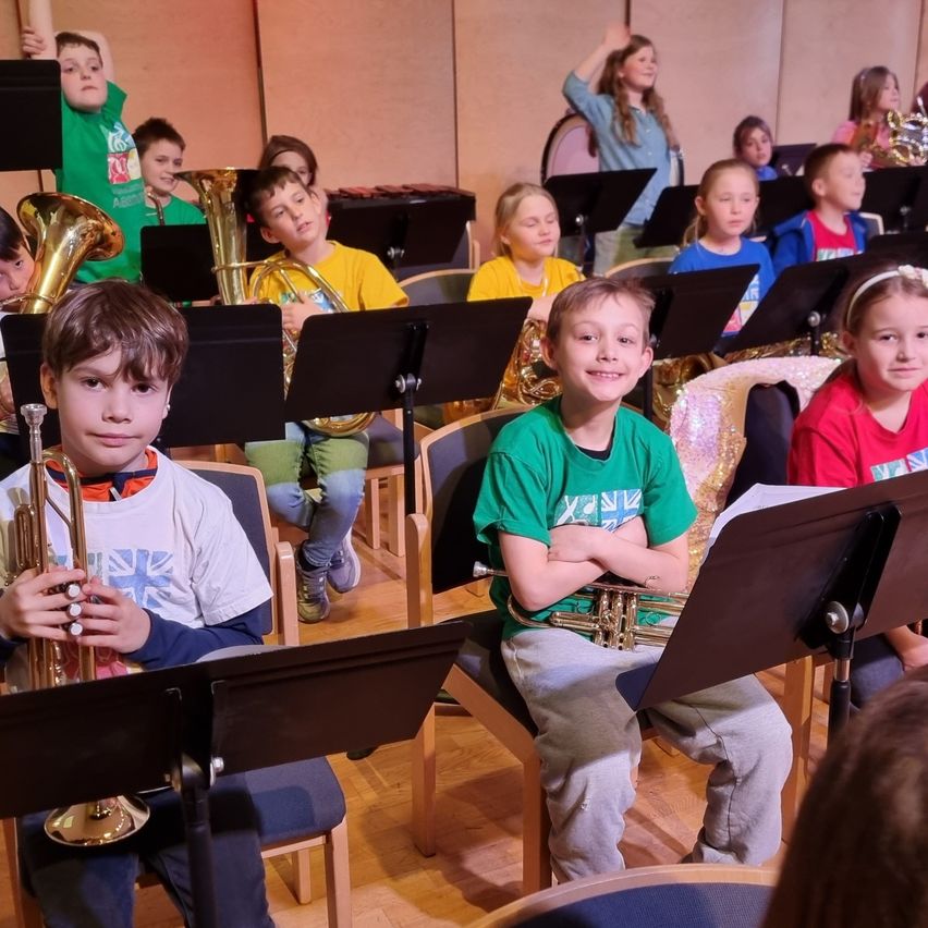Ein Klassenzimmer voller Kinder, einige sitzend und andere stehend, die alle verschiedene Musikinstrumente wie Trompeten und Posaunen spielen. Sie scheinen für eine Aufführung zu proben.