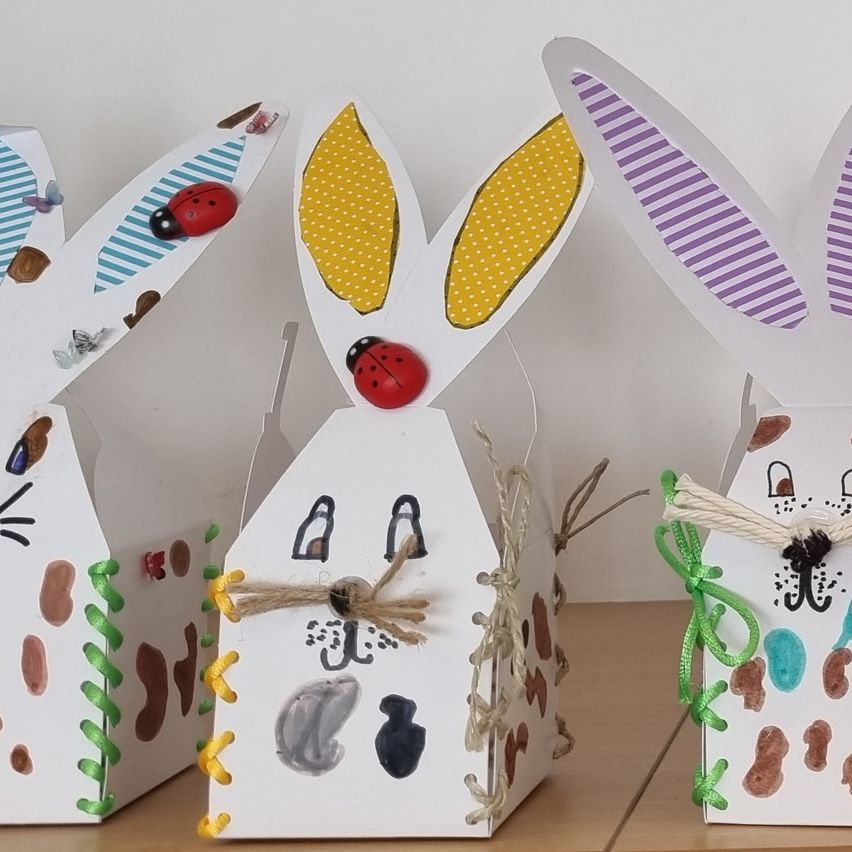 Vier dekorative Osterhasen aus weißem Papier mit bunten Details sind ausgestellt. Jeder Hase hat große Ohren, eine Marienkäfer und einzigartige Muster. Sie sind mit Bändern geschmückt und sitzen auf einer Holzoberfläche.