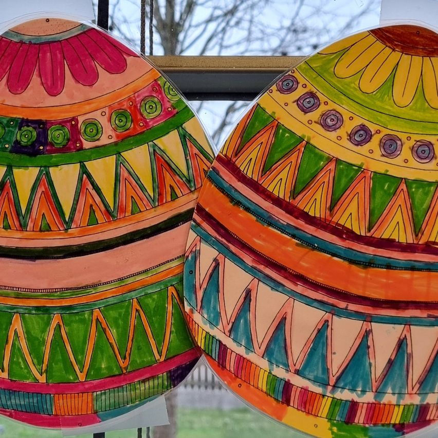 Zwei farbenfrohe Eier mit detaillierten Mustern und Designs werden vor einem Fenster ausgestellt. Ein Ei hat oben eine Sonnenblume, das andere ein Blumendesign.