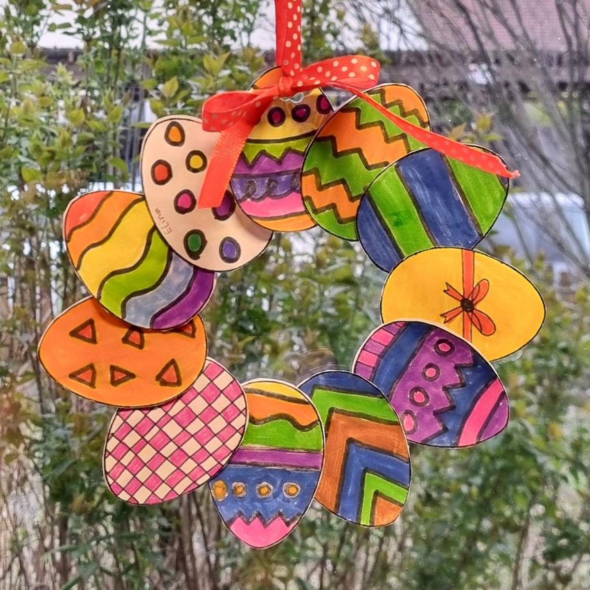 Ein farbenfroher Ostereier-Kranz mit verschiedenen Designs, der vor einem Fenster hängt. Oben ist ein rotes Band befestigt.