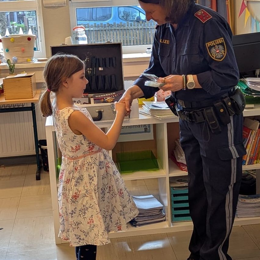 Eine Polizistin gibt einem jungen Mädchen einen kleinen Gegenstand in einem Klassenzimmer. Das Mädchen trägt ein Blumenkleid.