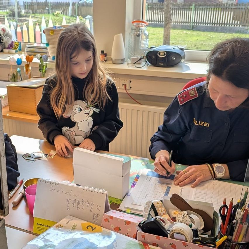 Ein Mädchen sitzt mit einem Notizbuch am Schreibtisch, während eine Polizistin auf einem Dokument schreibt. Auf dem Schreibtisch befinden sich verschiedene Gegenstände, darunter ein Stift, Scheren und eine kleine Schachtel. Die Szene spielt sich in einer Innenumgebung mit einem Fenster im Hintergrund ab.