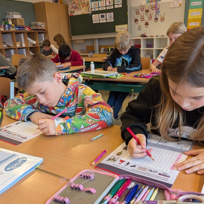 Schüler in einem Klassenzimmer beschäftigen sich mit Schreibaufgaben. Ein Junge konzentriert sich auf seinen Arbeitsbogen, während ein Mädchen neben ihm auf einem Kreuzworträtsel schreibt.