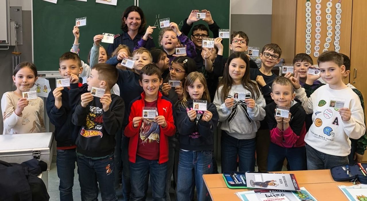Eine Gruppe von Kindern und Erwachsenen hält Karten hoch, mit einer Frau in Uniform vor einer Tafel im Klassenzimmer.