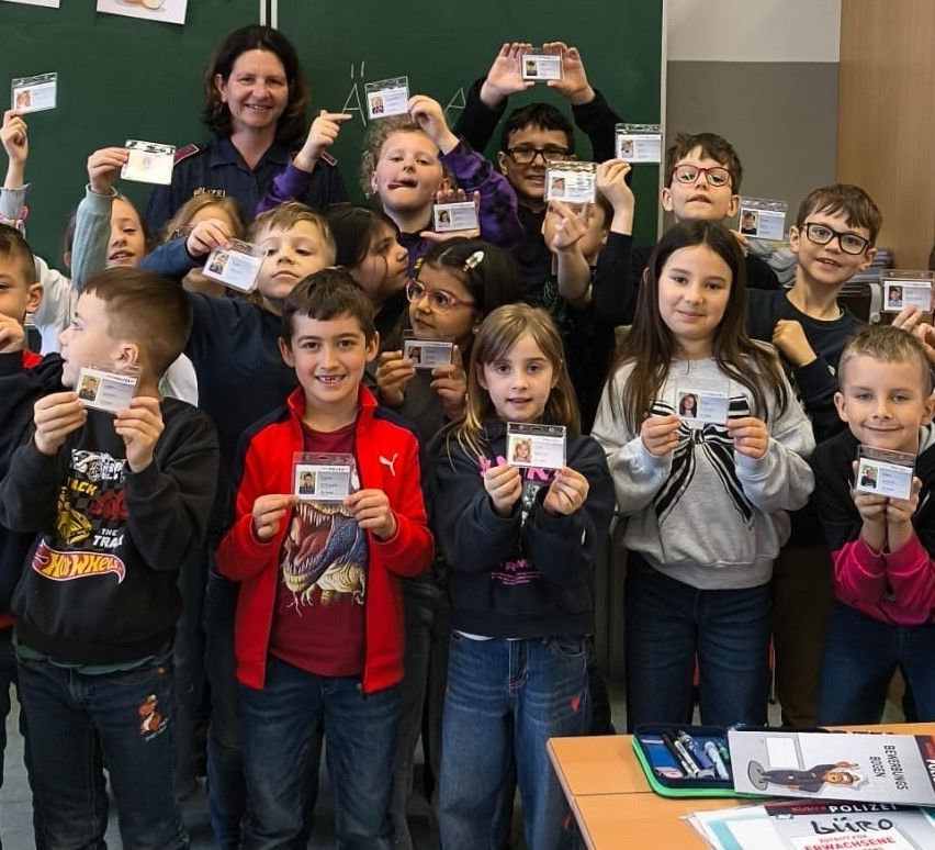 Eine Gruppe von Kindern und Erwachsenen hält Karten hoch, mit einer Frau in Uniform vor einer Tafel im Klassenzimmer.