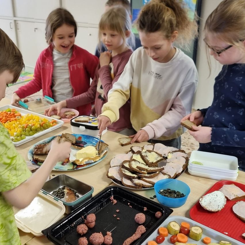 Kinder versammeln sich um einen Tisch und bereiten Sandwiches zu. Teller mit verschiedenen Lebensmitteln, wie Gemüse und Fleisch, sind vor ihnen platziert. Ein Junge in einem grünen Shirt schneidet ein Stück Fleisch, während andere ihre Sandwiches zubereiten.