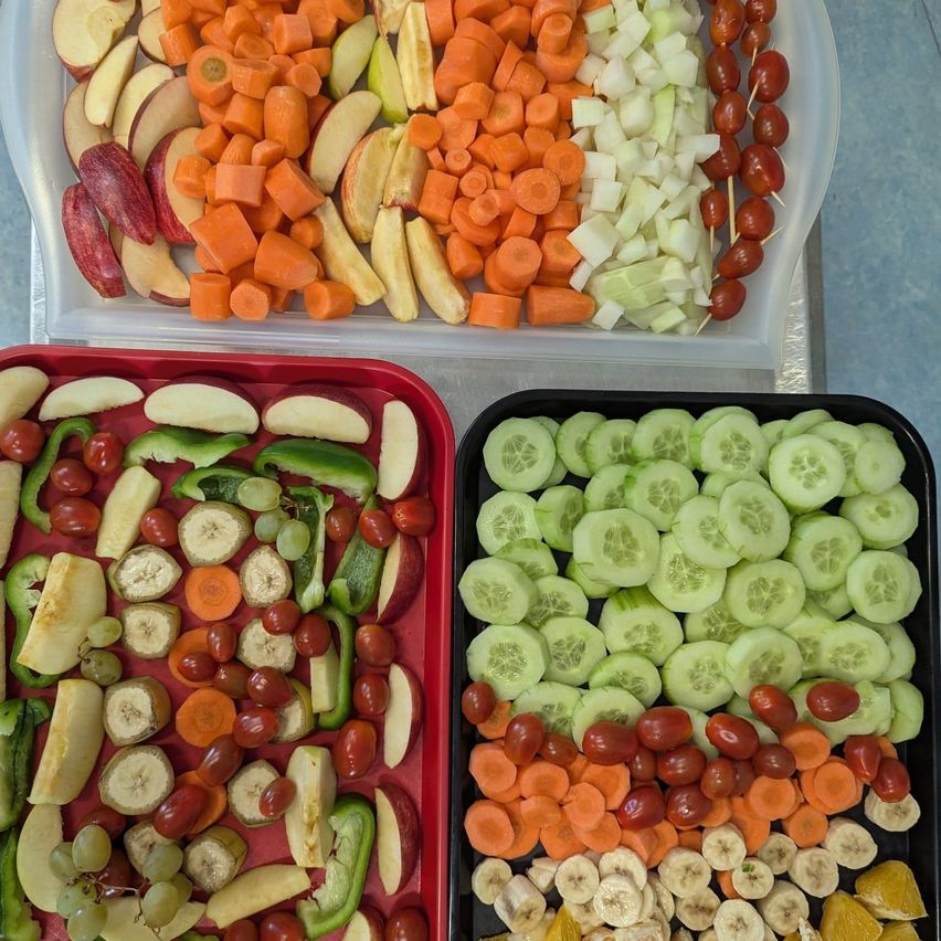 Drei Tabletts mit einer Vielfalt an geschnittenem Gemüse und Obst. Das oberste Tablett hat Karotten, Äpfel und Kirschtomaten. Das mittlere Tablett hat Paprika, Äpfel und Trauben. Das unterste Tablett hat Gurken, Karotten und Kirschtomaten.