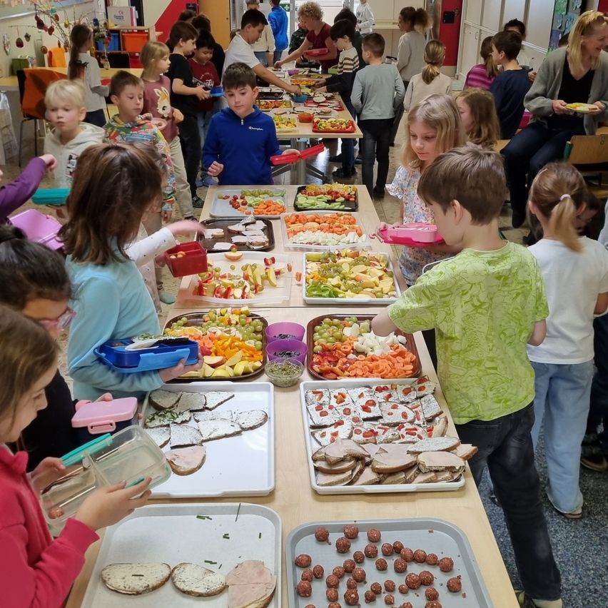 Ein Klassenzimmer voller Kinder, die sich um einen langen Tisch mit verschiedenen Lebensmitteln scharen. Jedes Kind hält eine Schale oder einen Behälter. Das Essen umfasst Obst, Sandwiches und andere Snacks.