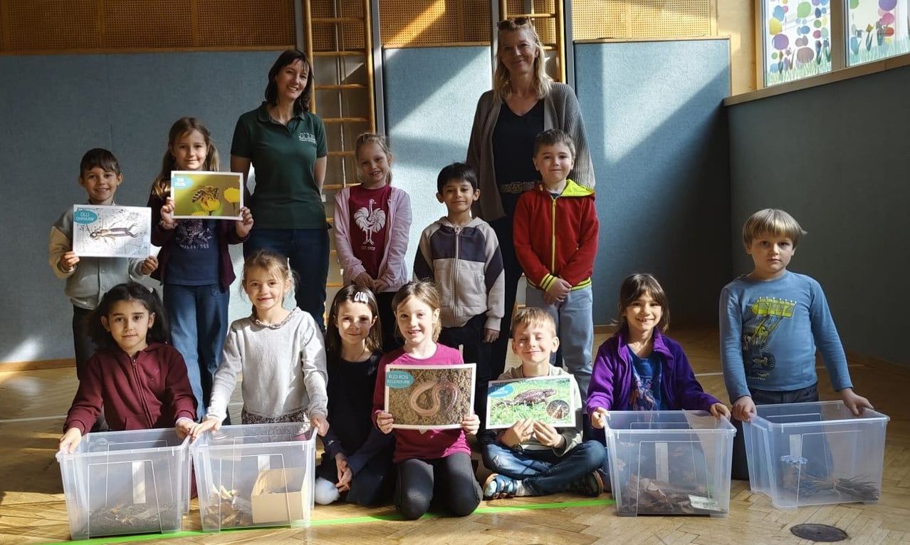 Eine Gruppe von Kindern in einem Klassenzimmer, die für ein Foto posieren. Sie halten Bilder von Tieren hoch. Eine Frau in Grün steht hinter ihnen.