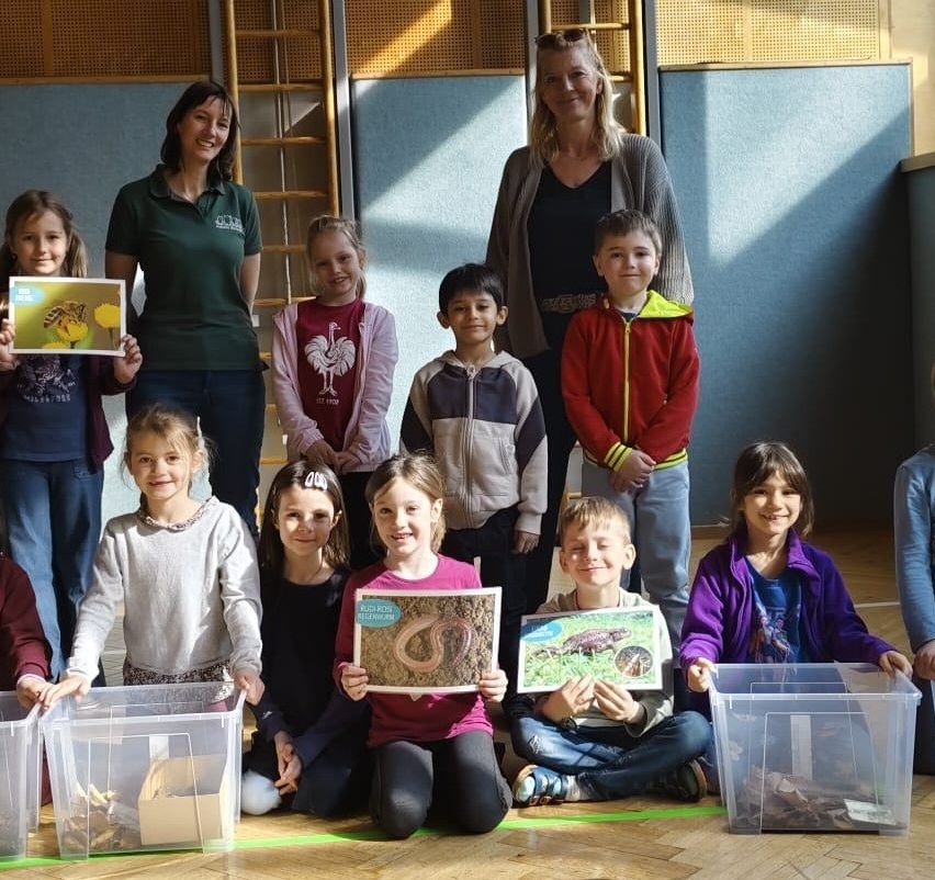 Eine Gruppe von Kindern in einem Klassenzimmer, die für ein Foto posieren. Sie halten Bilder von Tieren hoch. Eine Frau in Grün steht hinter ihnen.
