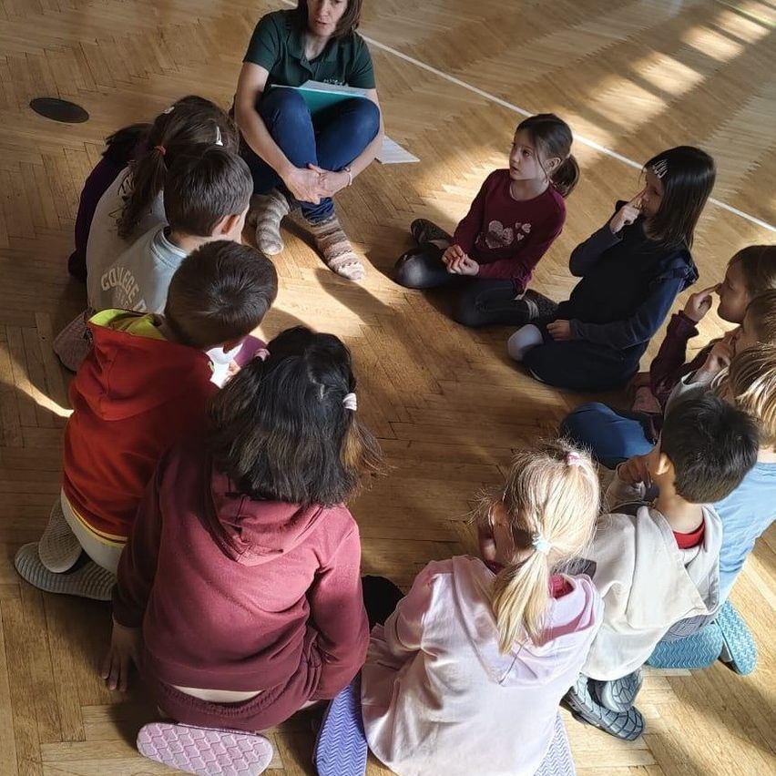 Eine Gruppe von Kindern sitzt im Kreis auf einem Holzboden und hört einer Frau mit einem Notizbuch zu. Die Kinder schauen aufmerksam auf die Frau.