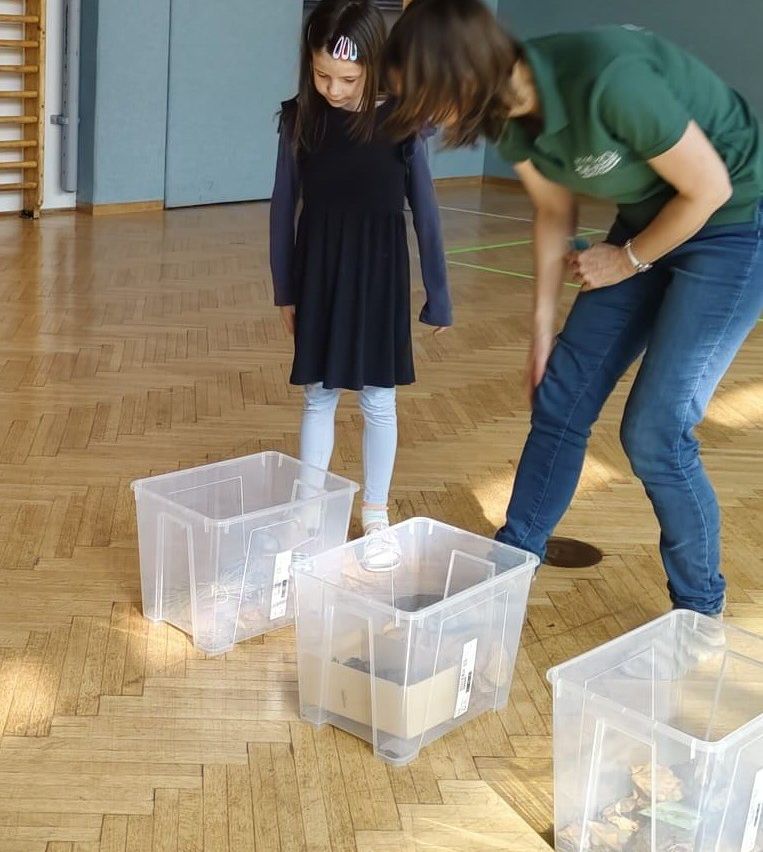 Eine Frau und ein junges Mädchen stehen auf einem Holzboden mit durchsichtigen Plastikbehältern. Die Frau beugt sich nach unten, um etwas aufzuheben, während das Mädchen mit den Händen hinter dem Rücken steht.