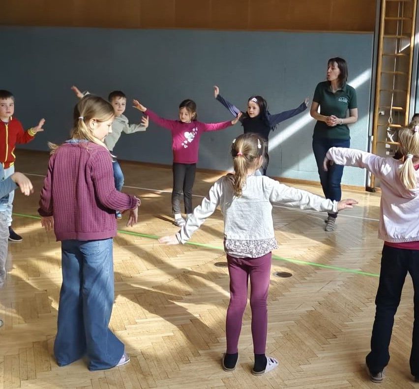 Eine Gruppe von Kindern übt in einem Raum Tanzschritte, während eine Frau sie beobachtet. Die Kinder haben ihre Arme erhoben und tragen verschiedenfarbige Kleidung.