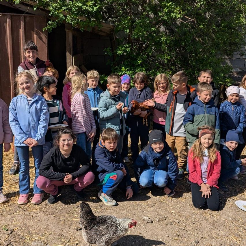 Eine Gruppe von Kindern und Erwachsenen posiert vor einer Scheune mit einem Huhn auf dem Boden für ein Foto. Einige Kinder halten Hühner.