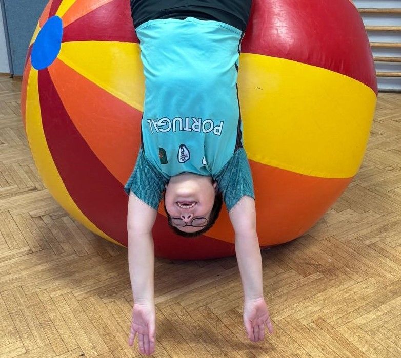 Ein junger Junge ist auf einem riesigen Strandball in einer Turnhalle auf dem Kopf stehend und trägt eine Brille.