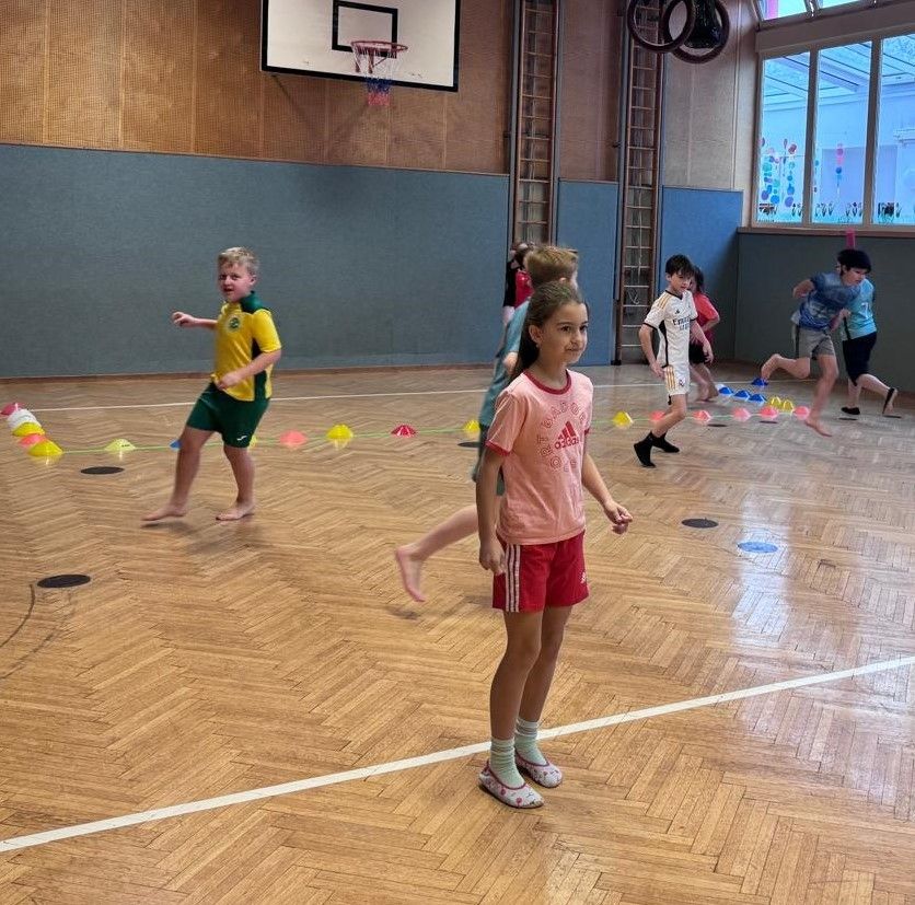 Mehrere Kinder spielen auf einem Hallenboden mit bunten Kegeln. Ein Kind läuft, während andere stehen. Der Boden hat weiße Markierungen und ein Basketballkorb ist an der Wand montiert.