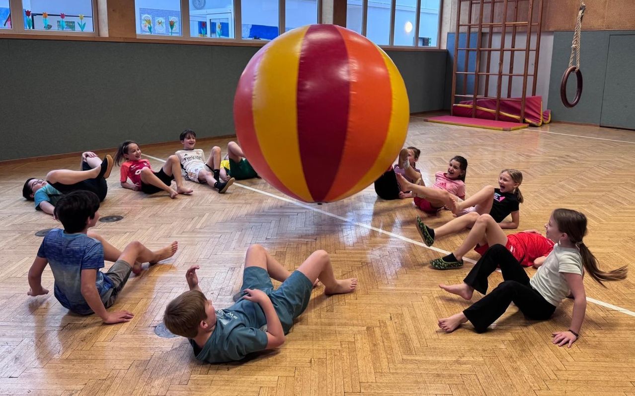 Kinder spielen mit einem großen mehrfarbigen Ball in einer Turnhalle, einige sitzen und andere liegen und lächeln.
