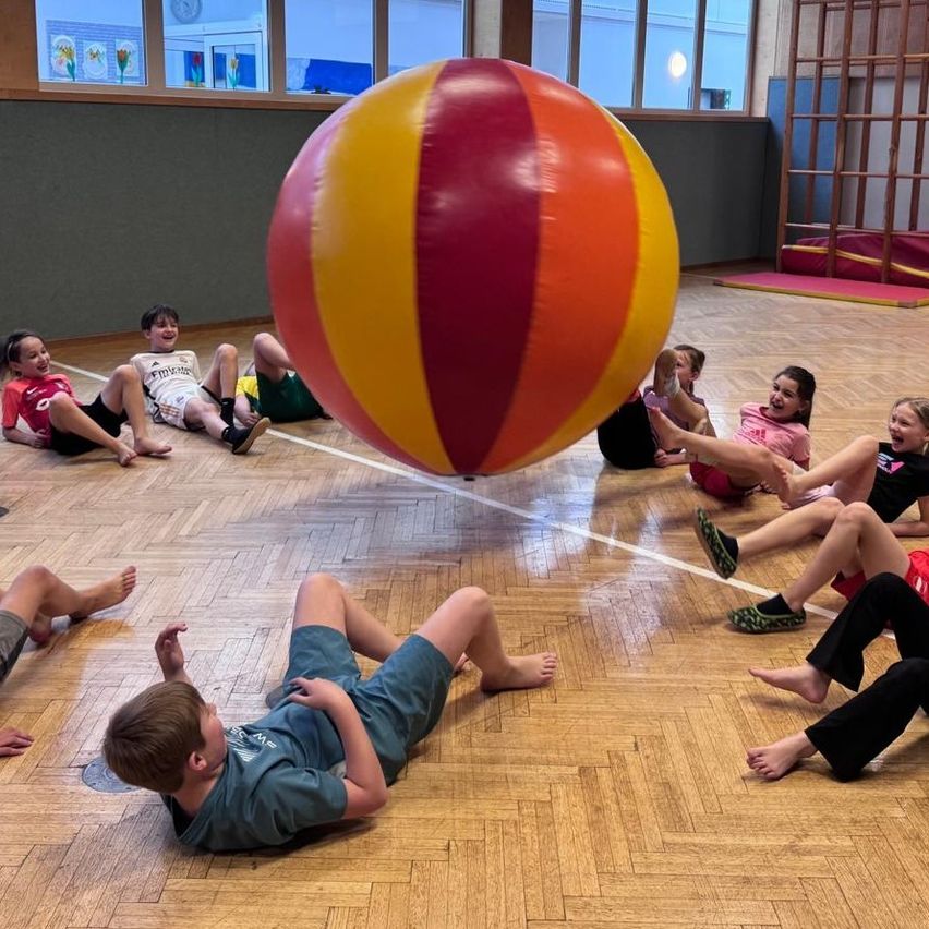 Kinder spielen mit einem großen mehrfarbigen Ball in einer Turnhalle, einige sitzen und andere liegen und lächeln.
