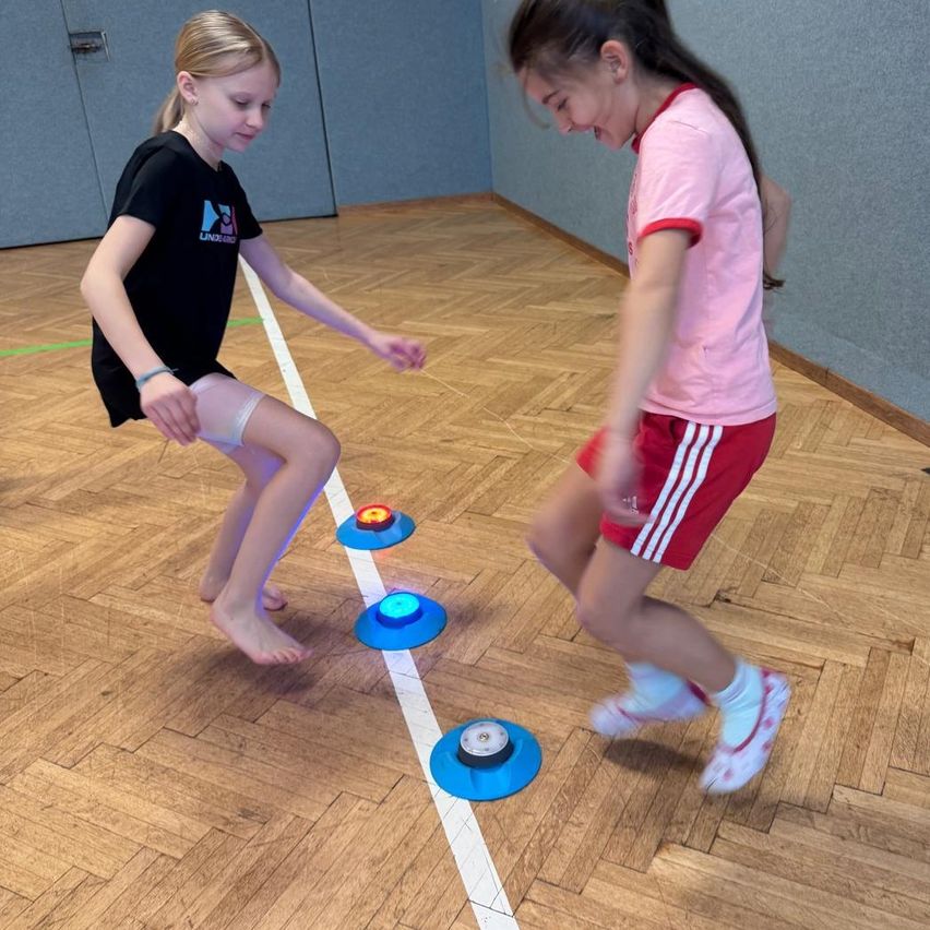Zwei Mädchen spielen mit Frisbees auf einem Holzboden, eines in einem schwarzen Shirt und das andere in einem rosa Shirt. Sie springen über farbige Scheiben.