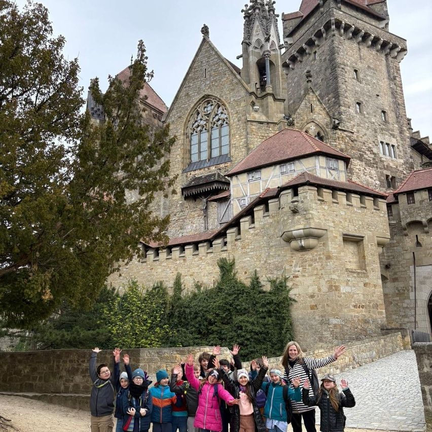 Eine Gruppe von Kindern und Erwachsenen posiert für ein Foto vor einer großen Burg. Sie alle lächeln und winken.