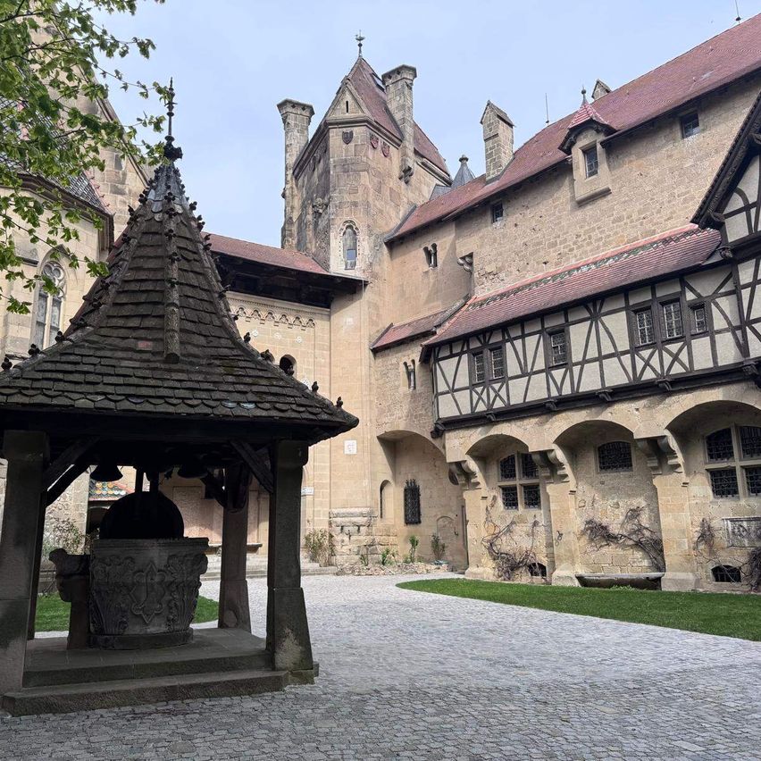 Ein historischer Schlosshof mit einem Steinbrunnen und einem großen Gebäude mit roten Dächern und Bogenfenstern.