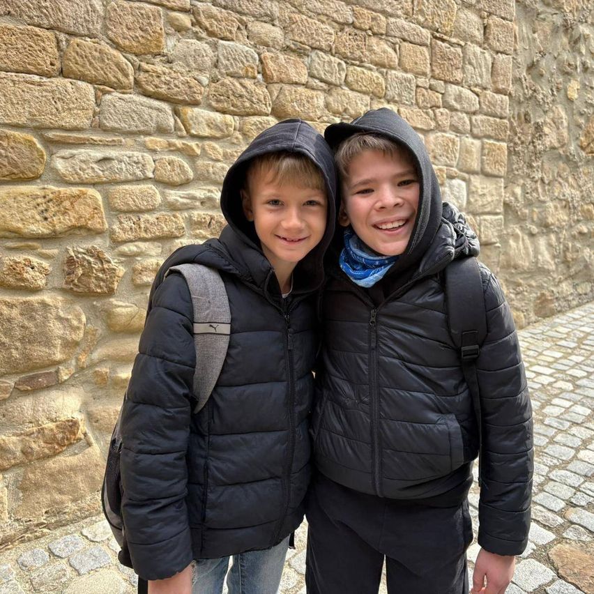 Zwei junge Jungen in schwarzen Jacken mit Kapuzen, lächelnd und eng nebeneinander stehend. Sie tragen beide Rucksäcke. Hinter ihnen ist eine Steinmauer.