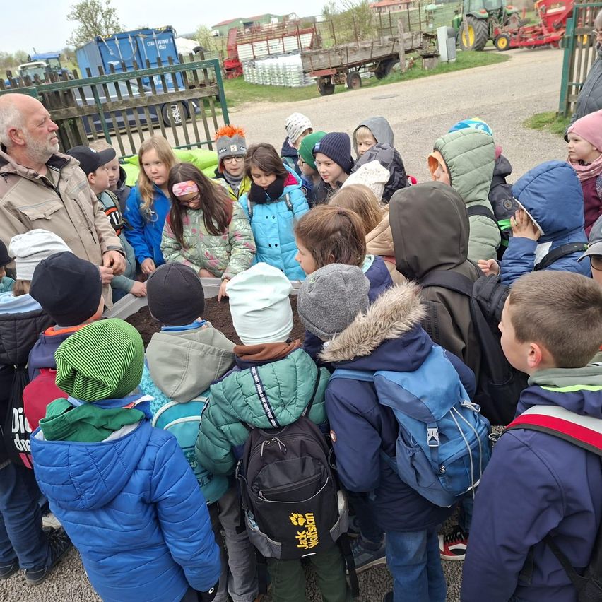 Eine Gruppe von Kindern und ein älterer Mann versammeln sich um einen Tisch im Freien, umgeben von verschiedenen Fahrzeugen und einem Zaun.