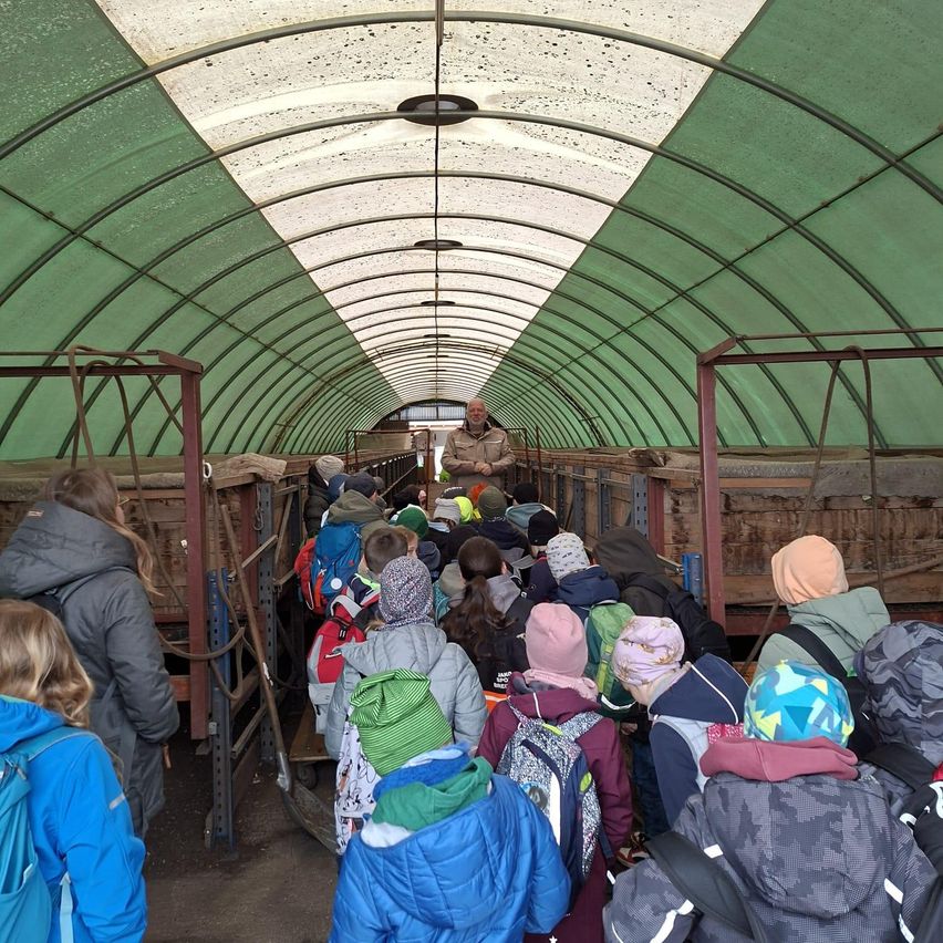 Eine Gruppe von Kindern in Winterkleidung steht in einem Tunnel und hört einem Mann in einer braunen Jacke zu. Der Tunnel ist mit grünen Wänden und Metallstrukturen ausgekleidet.