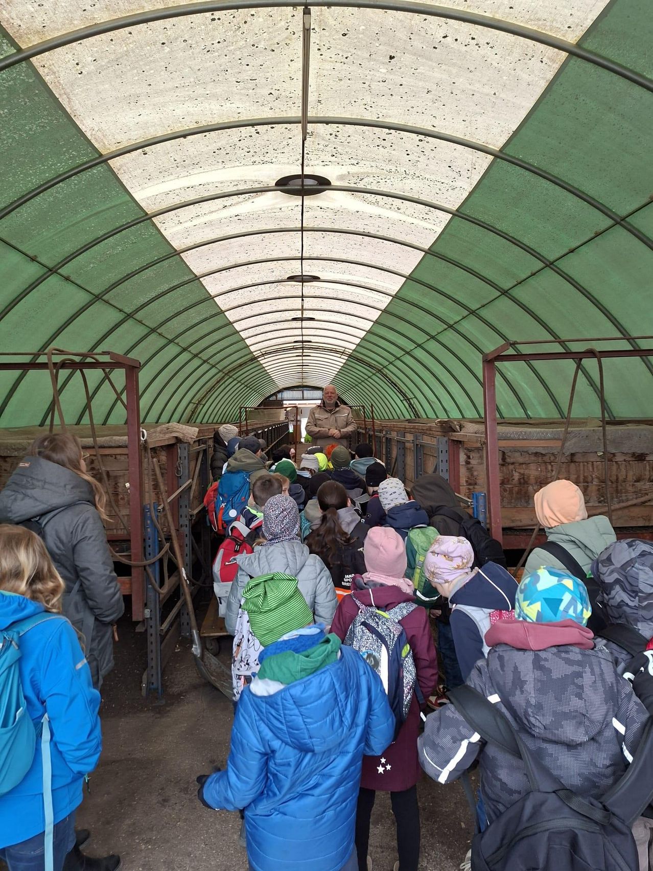 Eine Gruppe von Kindern in Winterkleidung steht in einem Tunnel und hört einem Mann in einer braunen Jacke zu. Der Tunnel ist mit grünen Wänden und Metallstrukturen ausgekleidet.