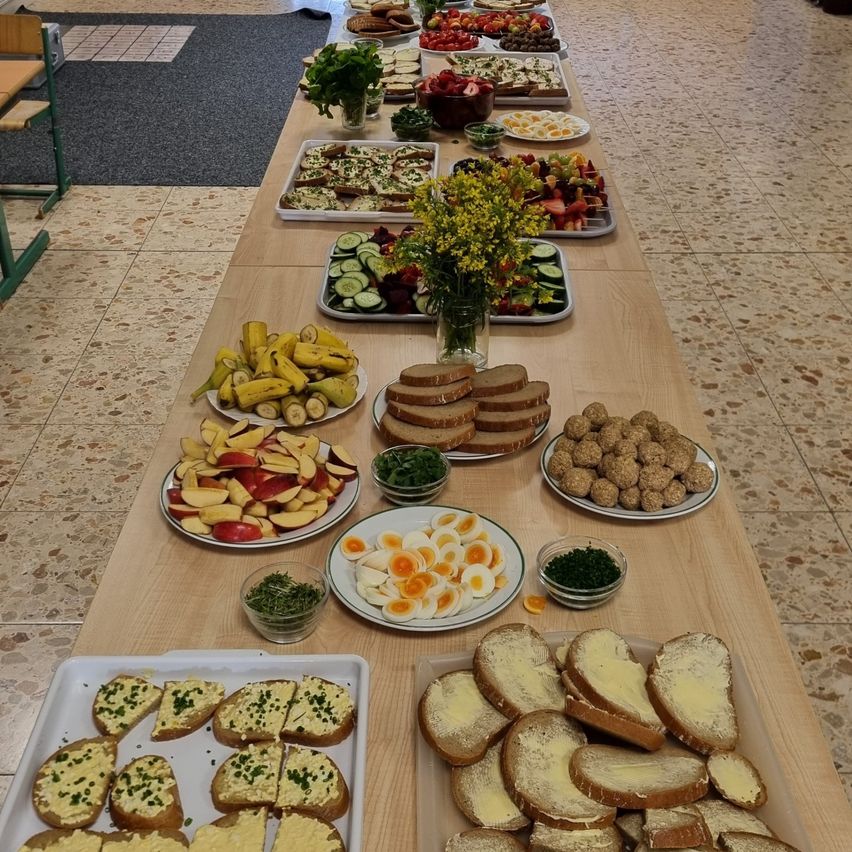 Ein langer Tisch, gefüllt mit verschiedenen Speisen wie Sandwiches, Obst, Gemüse und Desserts. Der Tisch ist drinnen mit einem gemusterten Boden aufgestellt.