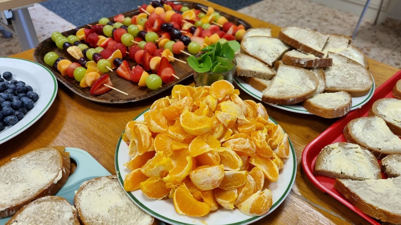 Ein Holztisch zeigt eine Vielzahl von Früchten. Ein Teller hat eine große Menge geschälter Orangen, während ein anderer Teller Obstspieße hat. Es gibt auch einen Teller mit Brot und ein kleines Glas mit Kräutern.