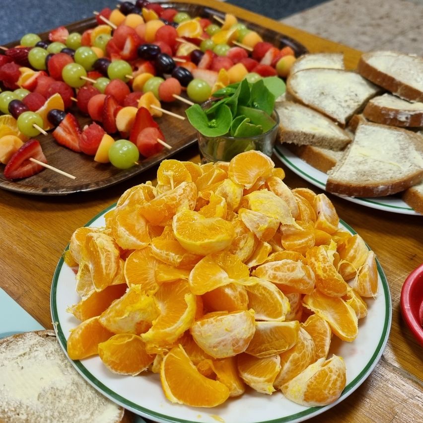 Ein Holztisch zeigt eine Vielzahl von Früchten. Ein Teller hat eine große Menge geschälter Orangen, während ein anderer Teller Obstspieße hat. Es gibt auch einen Teller mit Brot und ein kleines Glas mit Kräutern.