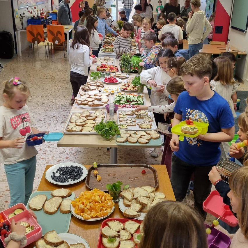 Ein Klassenzimmer mit Kindern, die um einen langen Tisch mit verschiedenen Speisen auf Tellern versammelt sind, darunter Sandwiches, Obst und Gemüse. Einige Kinder halten Lebensmittel in den Händen.