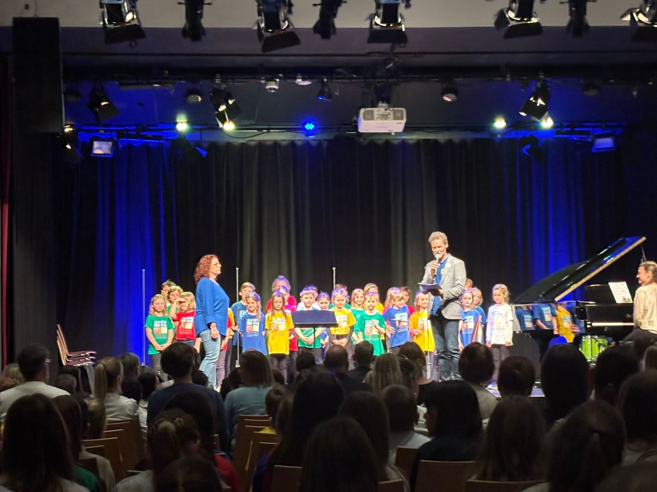 Ein Kinderchor in passenden gelben und grünen Shirts steht auf der Bühne vor einem Klavier mit einem Erwachsenen, der ein Mikrofon hält. Die Bühne ist mit blauer Beleuchtung ausgestattet.
