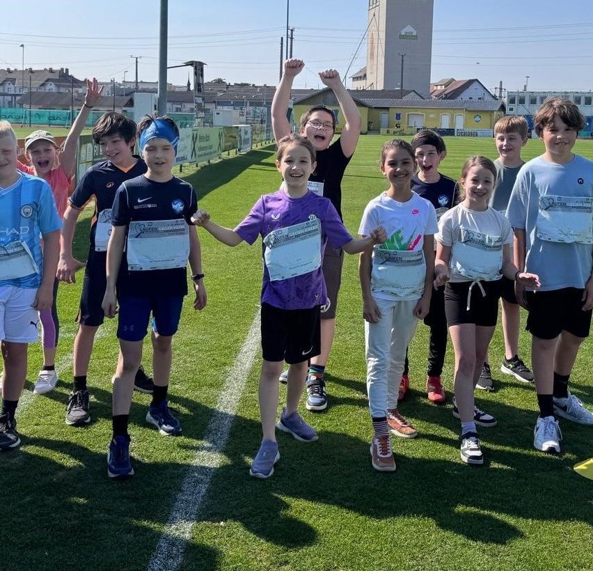 Eine Gruppe von Kindern mit Startnummern auf ihren Trikots steht auf einem Feld. Einer von ihnen hebt beide Hände. Sie tragen Turnschuhe und lächeln. Hinter ihnen befinden sich Häuser und ein Turm.