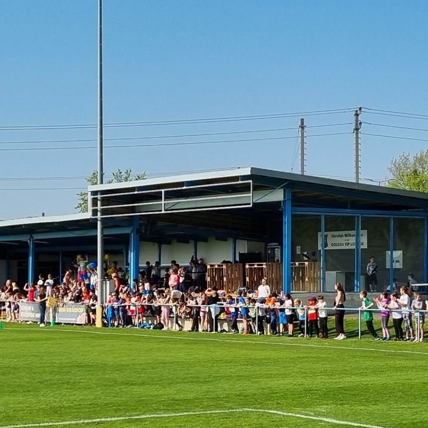 Eine Menschenmenge steht entlang des Zauns eines Sportfeldes. Ein großes Gebäude mit Glaswänden befindet sich hinter dem Zaun. Strommasten sind in der Ferne.